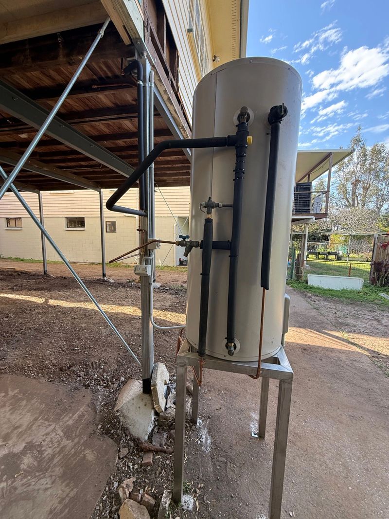 A tall cylindrical tank with black pipes and valves under a metal structure outdoors